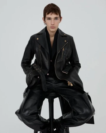 Man in black leather jacket and trousers poses while seated on a stool in a studio with bright, even lighting.