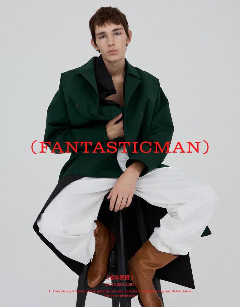 Editorial fashion shot of a young adult male in a green coat and white pants, sitting on a black stool against a white backdrop.