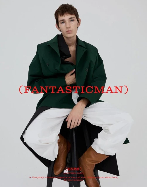 Editorial fashion shot of a young adult male in a green coat and white pants, sitting on a black stool against a white backdrop.