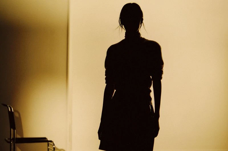 Silhouette of a woman in a dark sweater and skirt, standing indoors with a chair in the background, warm tones and studio lighting.
