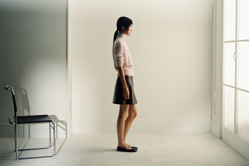 A slender woman in a pink sweater and brown skirt stands in profile in a bright white room with a chair and natural light.