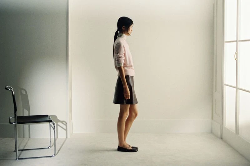 A slender woman in a pink sweater and brown skirt stands in profile in a bright white room with a chair and natural light.