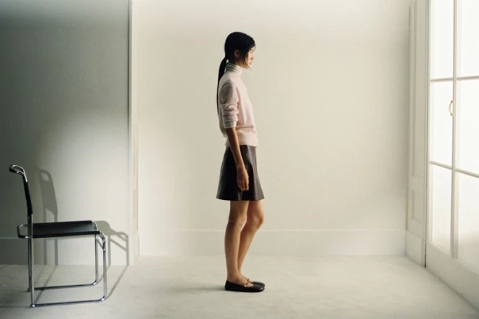 A slender woman in a pink sweater and brown skirt stands in profile in a bright white room with a chair and natural light.