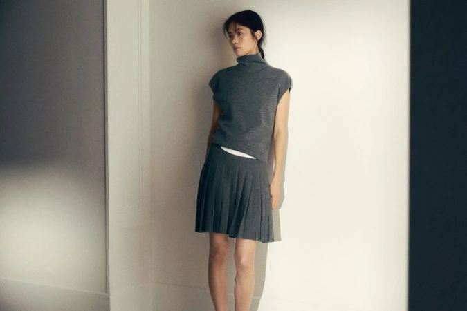 Woman poses in a grey sleeveless turtleneck and pleated skirt in a white corner studio, showcasing a minimalist fashion aesthetic.