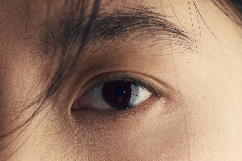 Close-up shot of an adult human eye with dark brown iris surrounded by skin and hair. Eye reflects a light source.