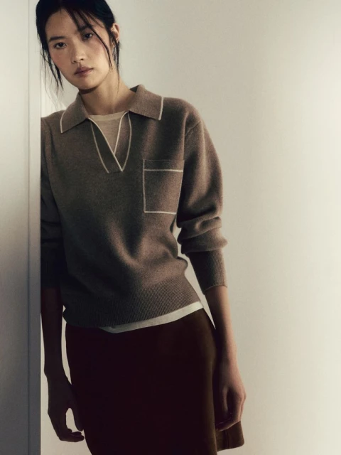 A young Asian woman in a brown knit polo shirt and dark skirt poses indoors against a light wall, offering a sophisticated, neutral-toned aesthetic.