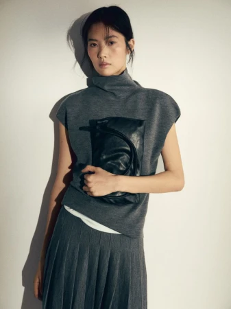 Studio shot of a female model wearing a grey sleeveless turtleneck top, pleated grey skirt, and holding a black clutch purse.