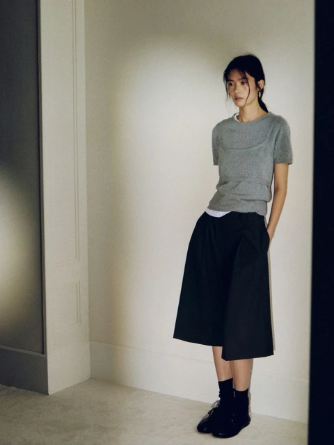 A young woman with dark hair in a gray short-sleeve top and black wide-leg pants poses in a softly lit interior against a cream-colored wall.