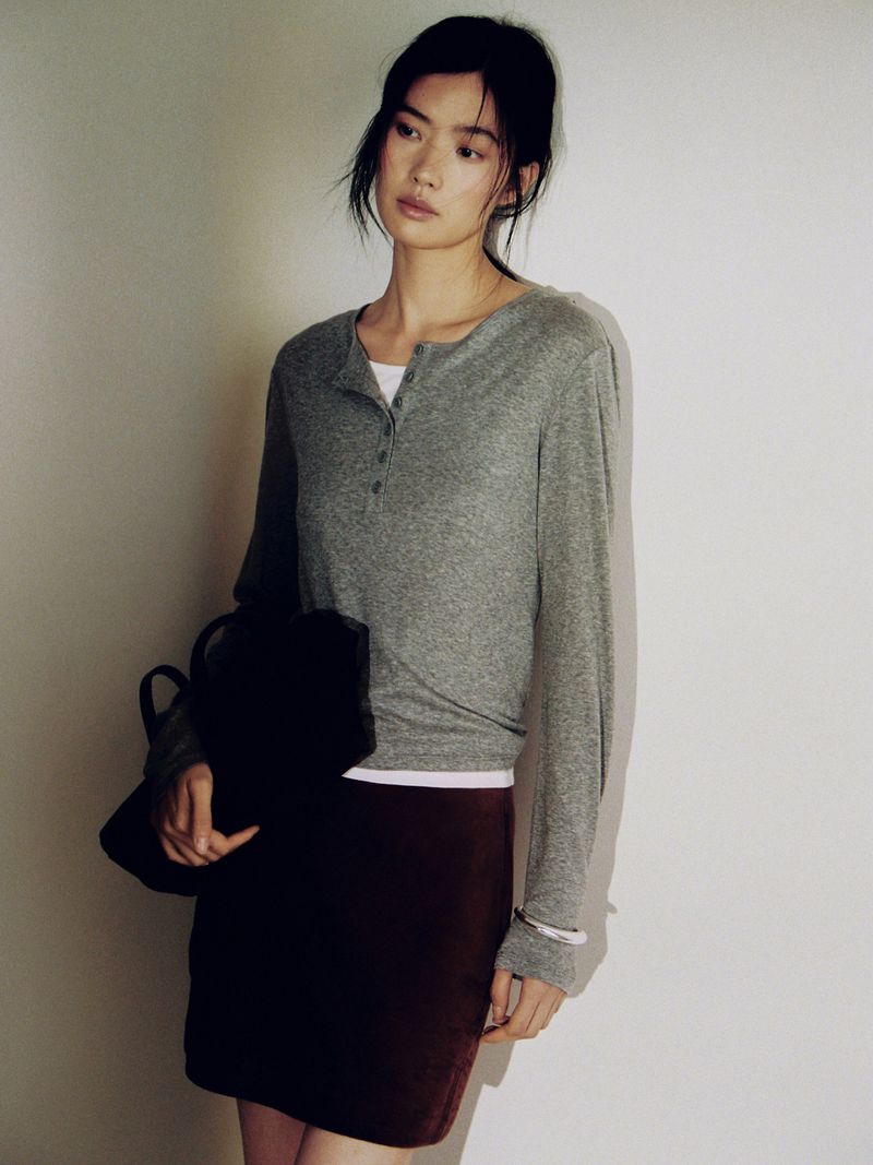 Studio shot of an Asian woman posing, wearing a grey long-sleeved shirt and a maroon skirt, holding a black bag.
