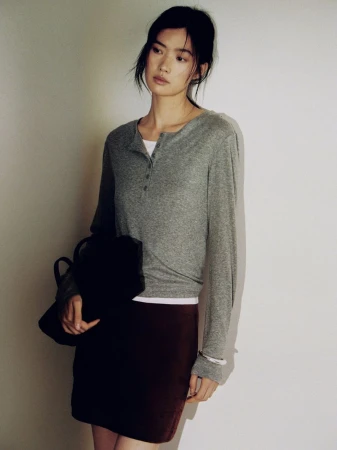 Studio shot of an Asian woman posing, wearing a grey long-sleeved shirt and a maroon skirt, holding a black bag.
