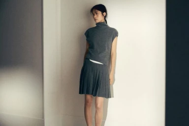 Model wearing a grey turtleneck top and pleated skirt leaning against a pale wall for Massimo Dutti
