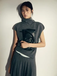 Model wearing a grey turtleneck top and pleated skirt holding a black leather clutch against a plain light wall