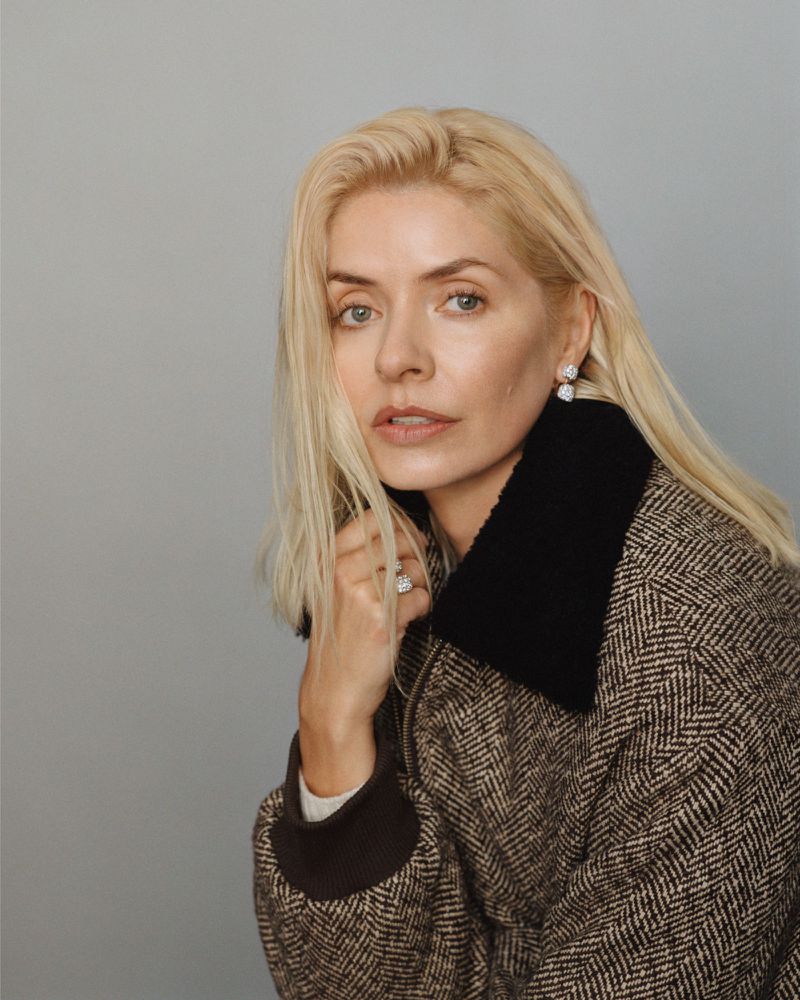 Portrait of a blond woman wearing a herringbone coat with a dark collar, gazing calmly at the camera in a studio setting with a plain gray background.