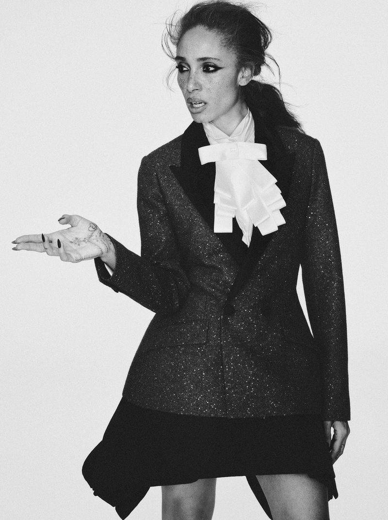 A woman poses in a blazer, blouse and skirt. The image is black and white with a white background.