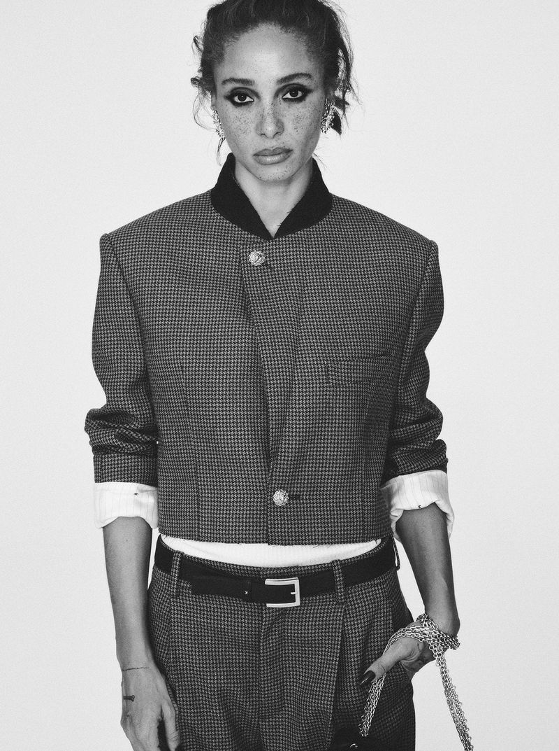 Black and white portrait of an adult woman wearing a cropped jacket, belt and pants. The model poses in front of a plain backdrop.