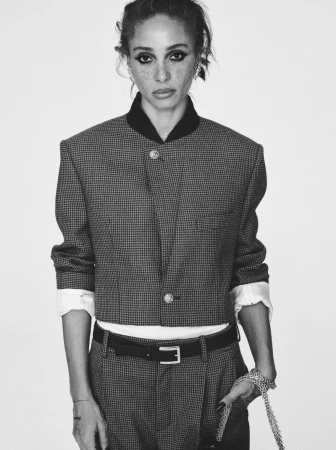 Black and white portrait of an adult woman wearing a cropped jacket, belt and pants. The model poses in front of a plain backdrop.