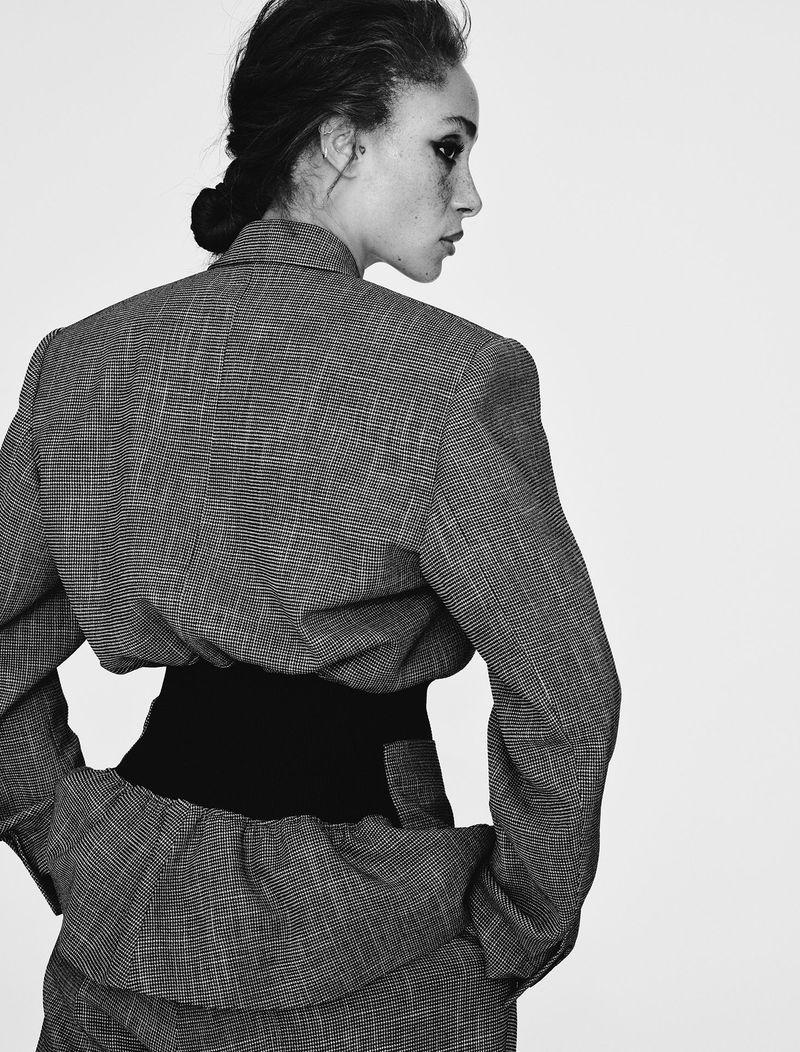 Black and white portrait of a woman in a grey patterned suit, turned to the side with dark eye makeup and a dark, cinched waist.