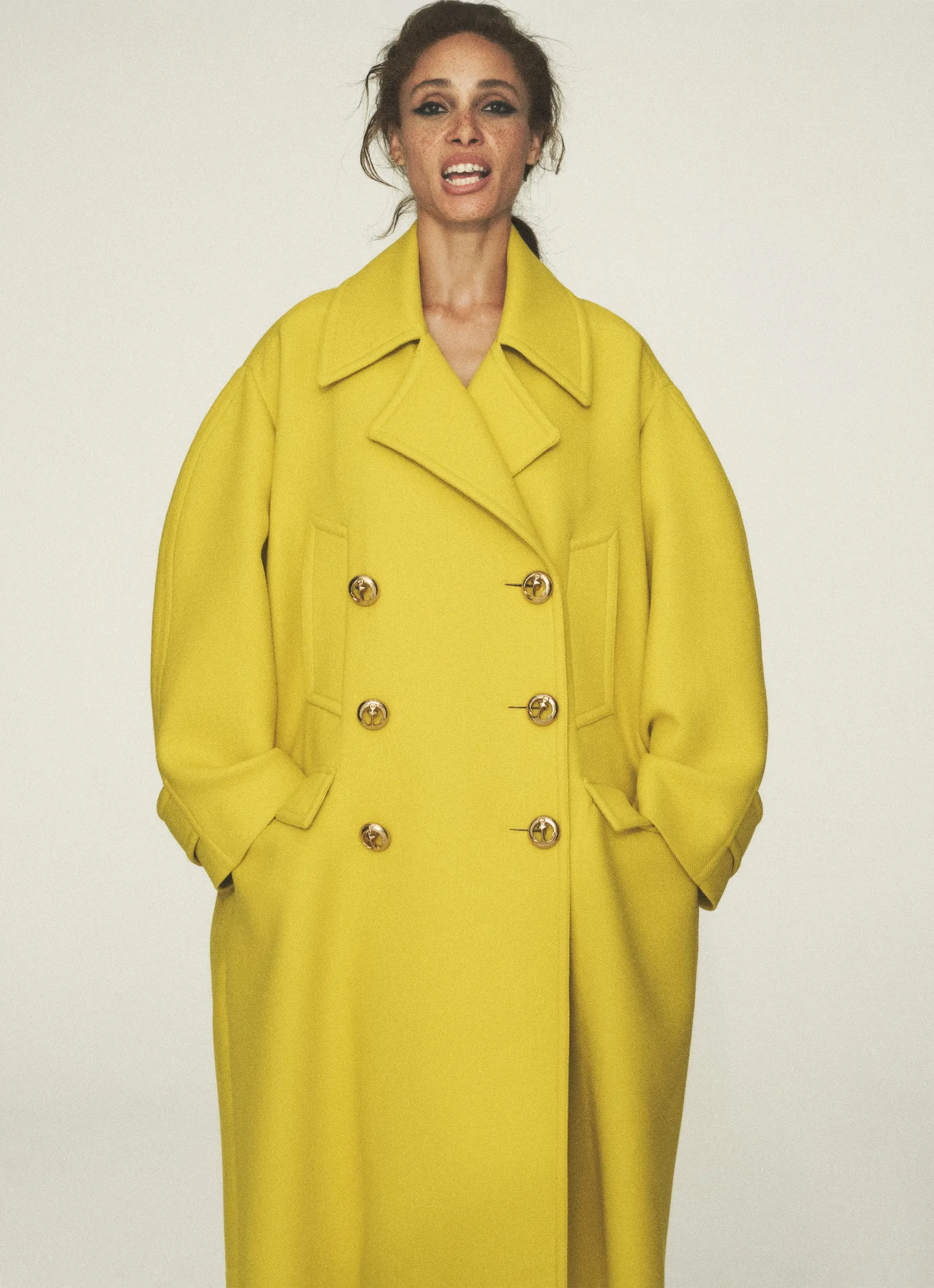 Woman in bright yellow double-breasted wool overcoat with gold buttons in a fashion studio shot against a clean white backdrop.