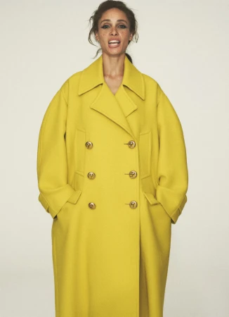 Woman in bright yellow double-breasted wool overcoat with gold buttons in a fashion studio shot against a clean white backdrop.