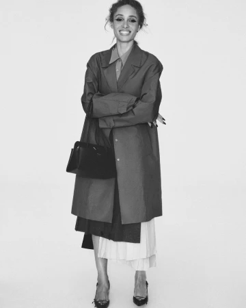 Model Adwoa Aboah standing with arms crossed wearing an oversized coat, pleated skirt, and heels against a white studio backdrop.
