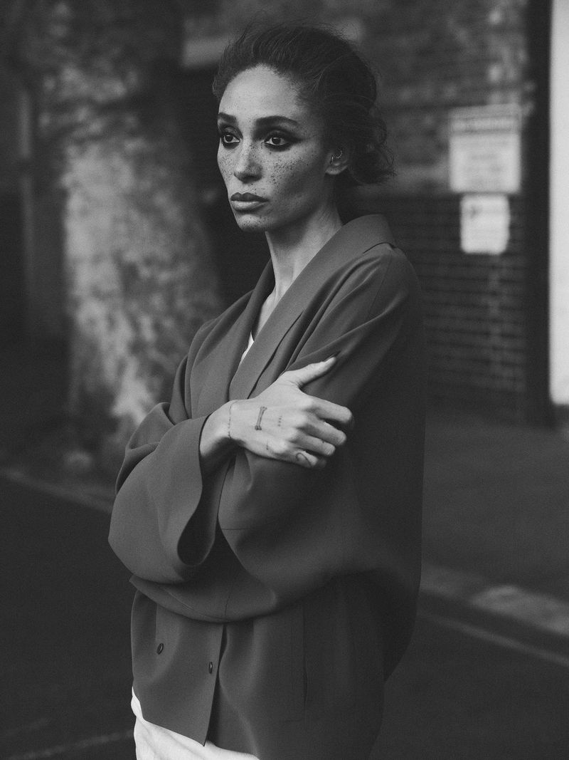 Black and white portrait of a woman in a red coat with arms crossed, standing outdoors on a city street with brick buildings.