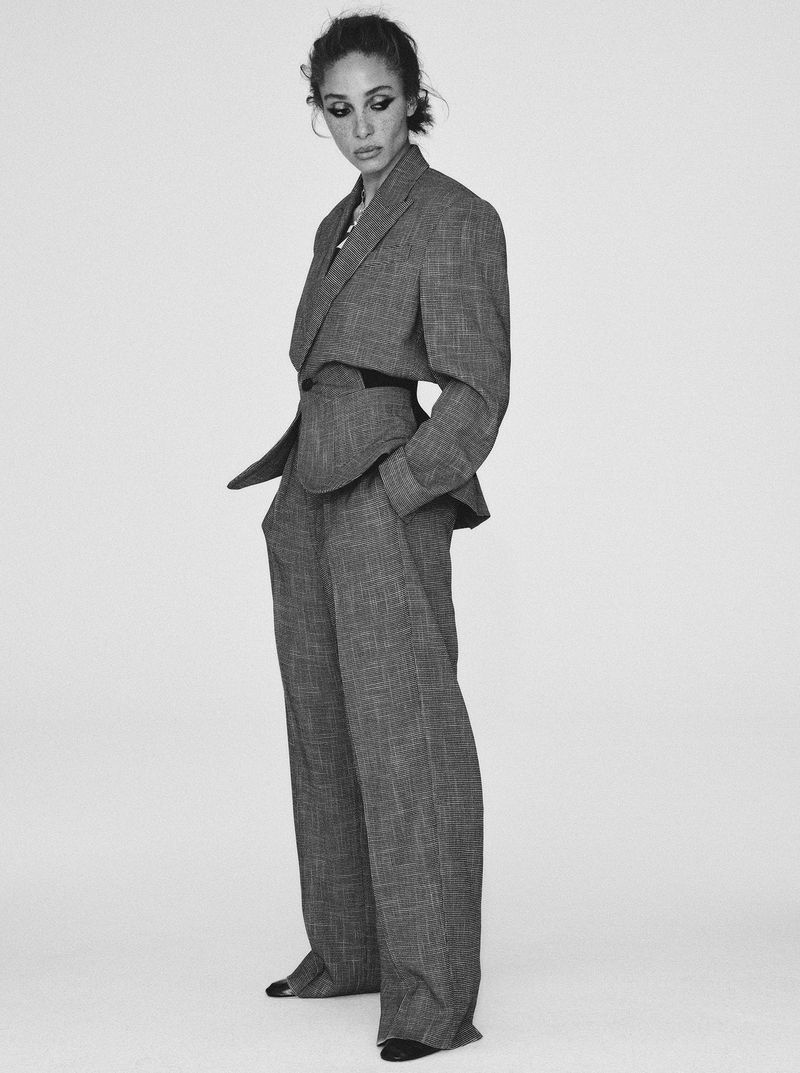 Monochrome portrait of a stylish adult woman in a patterned grey suit. She poses in a photo studio with hands in pockets.