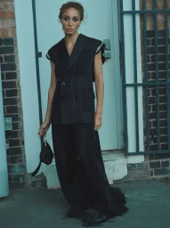 Model wearing a black sleeveless blazer and long trousers holding a handbag in front of a blue-toned door