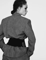 Adwoa Aboah seen from behind in a structured tweed suit with a cinched black waist panel against a plain white studio background.