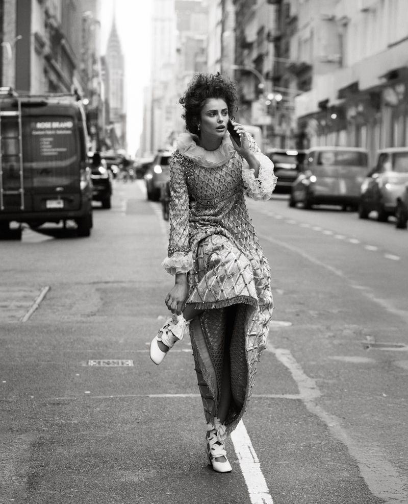 A female fashion model in an ornate dress walks on a city street while talking on a cell phone. Cars and buildings in the background.