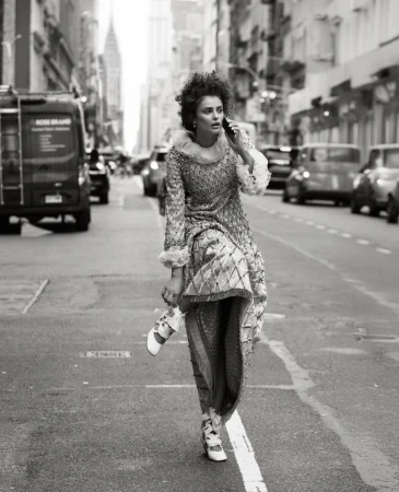 A female fashion model in an ornate dress walks on a city street while talking on a cell phone. Cars and buildings in the background.