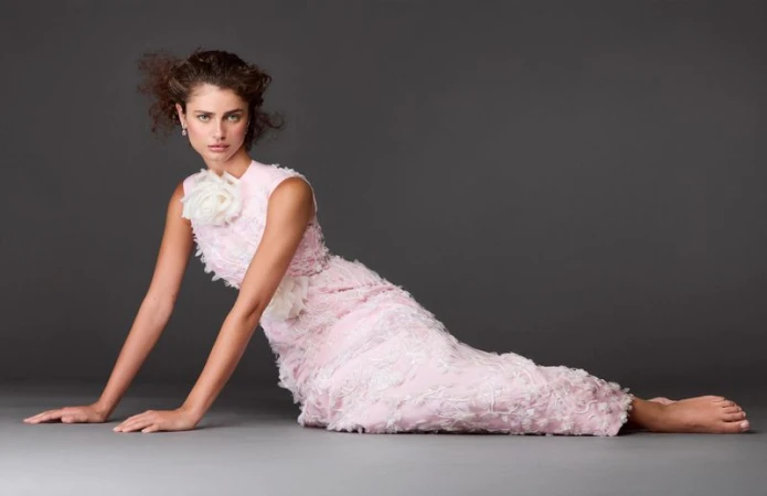 Woman in pink gown with white floral embellishments poses on gray studio backdrop, capturing elegant fashion and style.