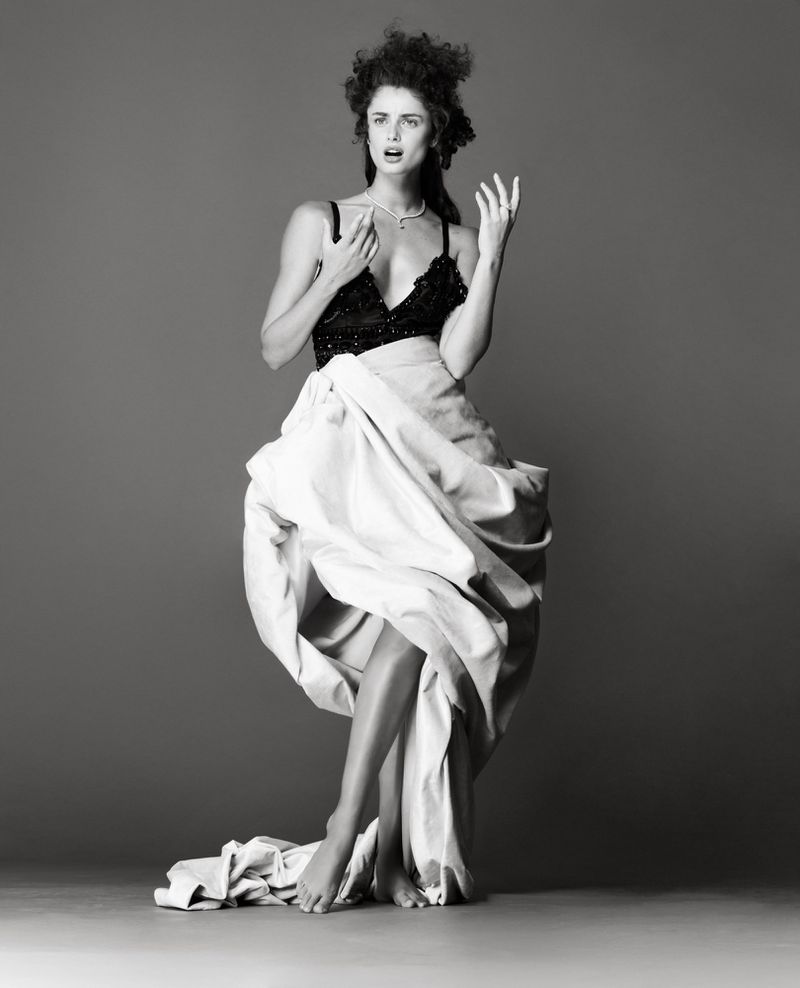 Black and white portrait of woman in a black bra and draped white fabric, striking a dramatic pose against a neutral gray backdrop.