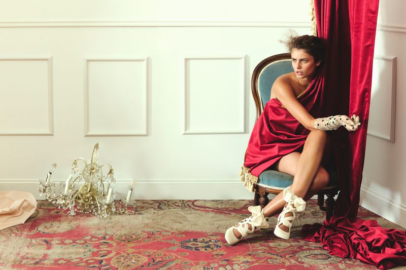 A woman sits draped in red fabric on an ornate chair in an indoor setting with a chandelier and red patterned carpet.