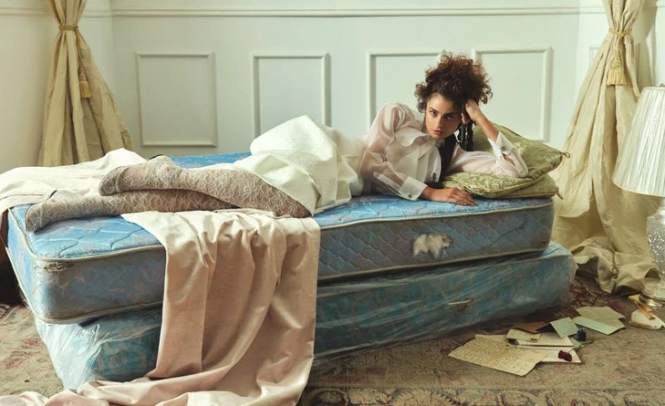 Woman with curly hair lies on an old mattress indoors. She wears a white top. Beige patterned stockings, vintage letters on the floor.