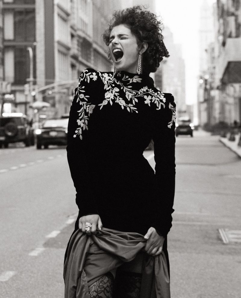 Black and white portrait of a curly-haired woman in an embroidered black dress, standing on a city street with cars and buildings.