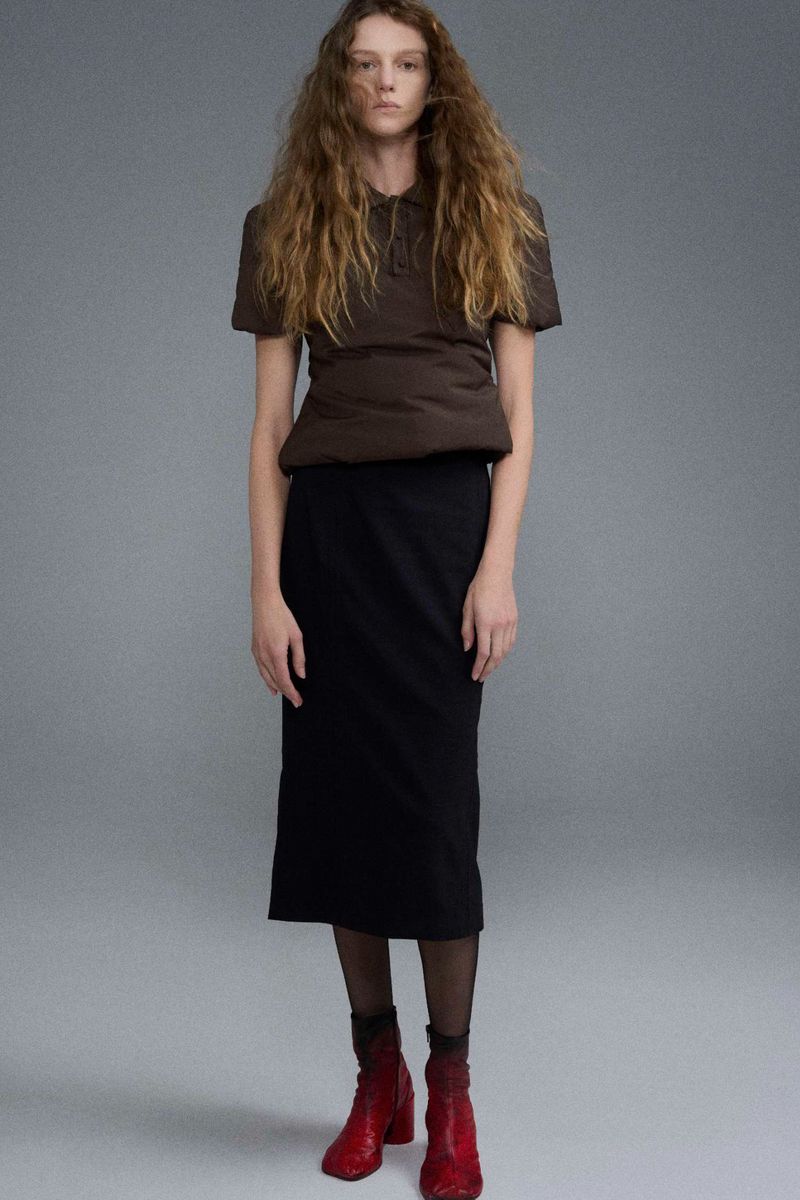 Fashion shot of a model wearing a brown top, black skirt, red boots, and sheer socks, posing in a photo studio with neutral background.