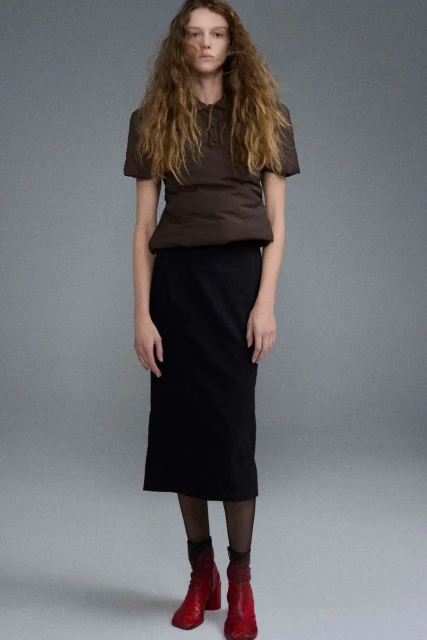 Fashion shot of a model wearing a brown top, black skirt, red boots, and sheer socks, posing in a photo studio with neutral background.