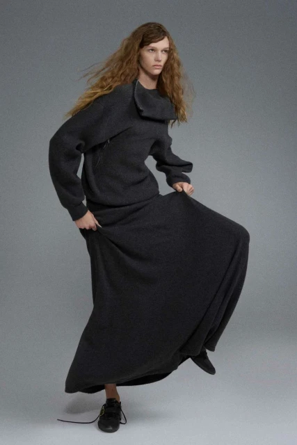 Fashion editorial shot of a woman in a dark grey, knitted, oversized sweater and long skirt, paired with black athletic shoes in studio.