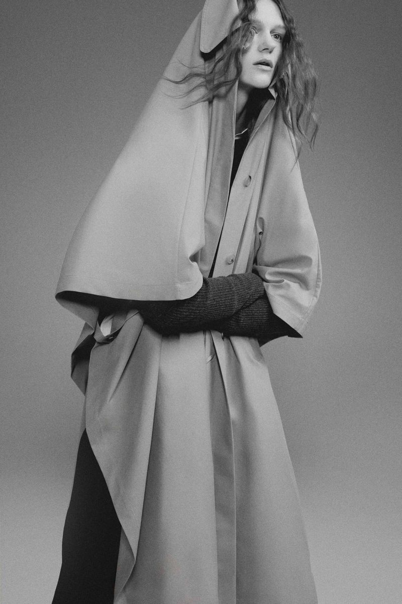 Monochrome portrait of a woman in a tan trench coat with a hood, styled with textured gloves and a neutral studio backdrop.