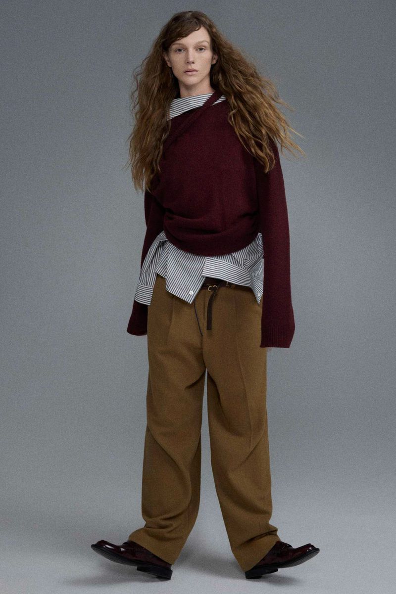 Fashion editorial shot of a young adult woman posing in a burgundy sweater, striped shirt, and khaki pants against a grey backdrop.