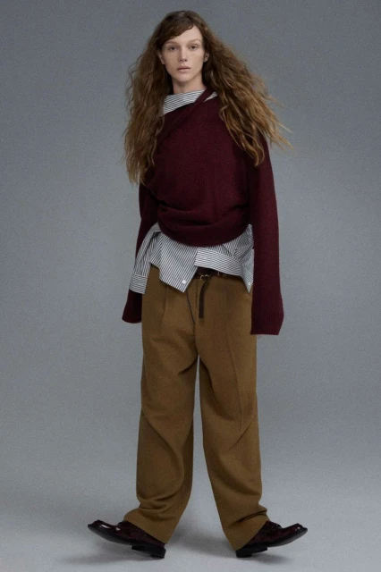 Fashion editorial shot of a young adult woman posing in a burgundy sweater, striped shirt, and khaki pants against a grey backdrop.
