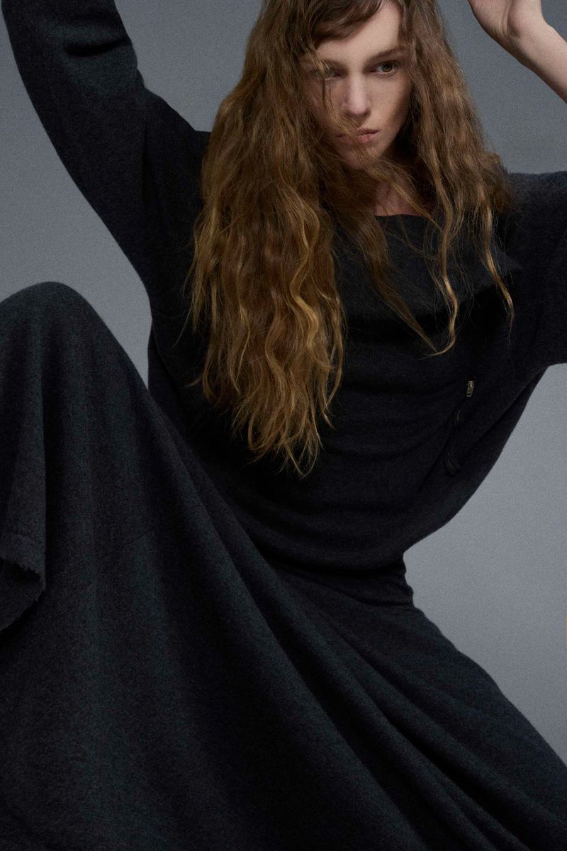 A stylish woman with long, wavy hair is shown in a medium shot, wearing a dark-colored dress or gown and seated against a neutral background.