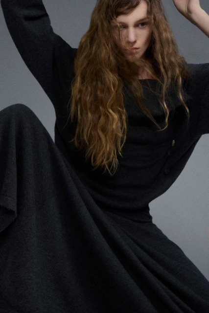 A stylish woman with long, wavy hair is shown in a medium shot, wearing a dark-colored dress or gown and seated against a neutral background.