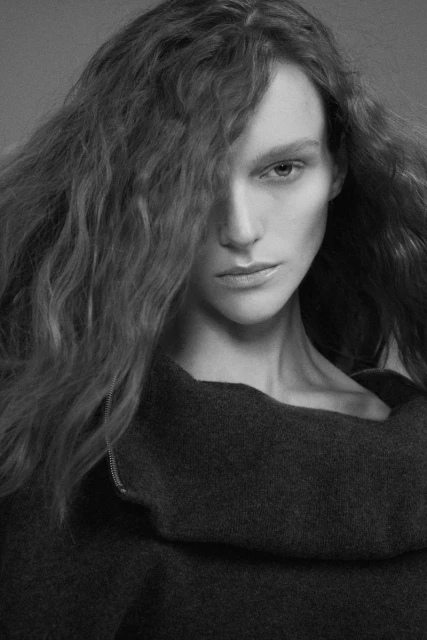 Monochrome portrait of a woman with wavy hair wearing a dark, cowl-neck top, shot indoors against a neutral gray background.