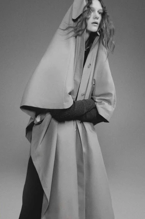 Model wearing an oversized hooded coat and dark long sleeves against a solid grey studio background in a fashion portrait.