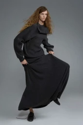 Model in a dark grey outfit with a long skirt and asymmetrical collar posing against a seamless grey studio background.
