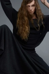 Model with long wavy brown hair wearing a dark grey textured knit dress against a neutral grey studio backdrop.
