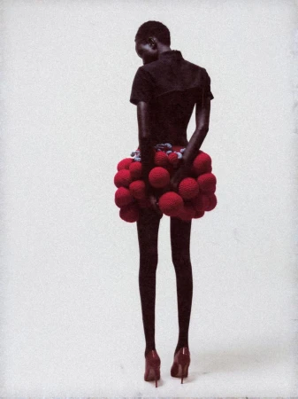 Back view of a slender African-American fashion model wearing a black top and a unique red pompom skirt with matching heels in a studio.