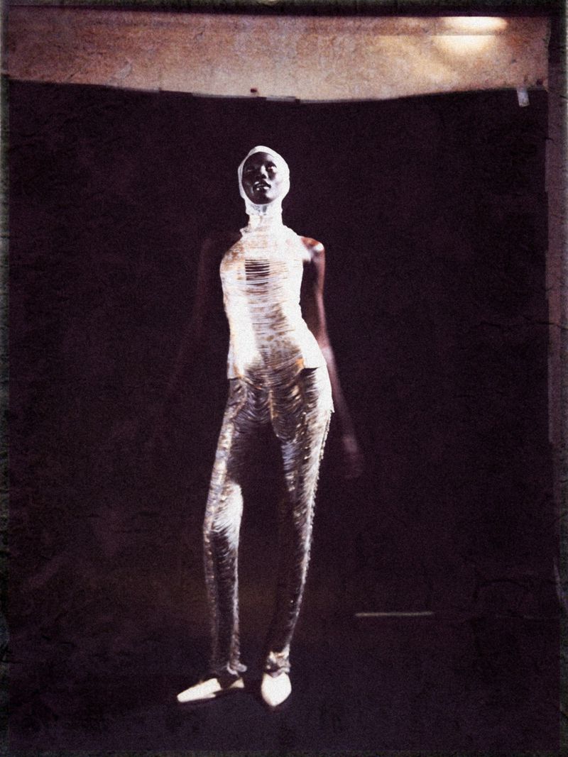 A slender Black female fashion model poses in a sleeveless metallic outfit against a shadowy backdrop, exuding confidence and style.