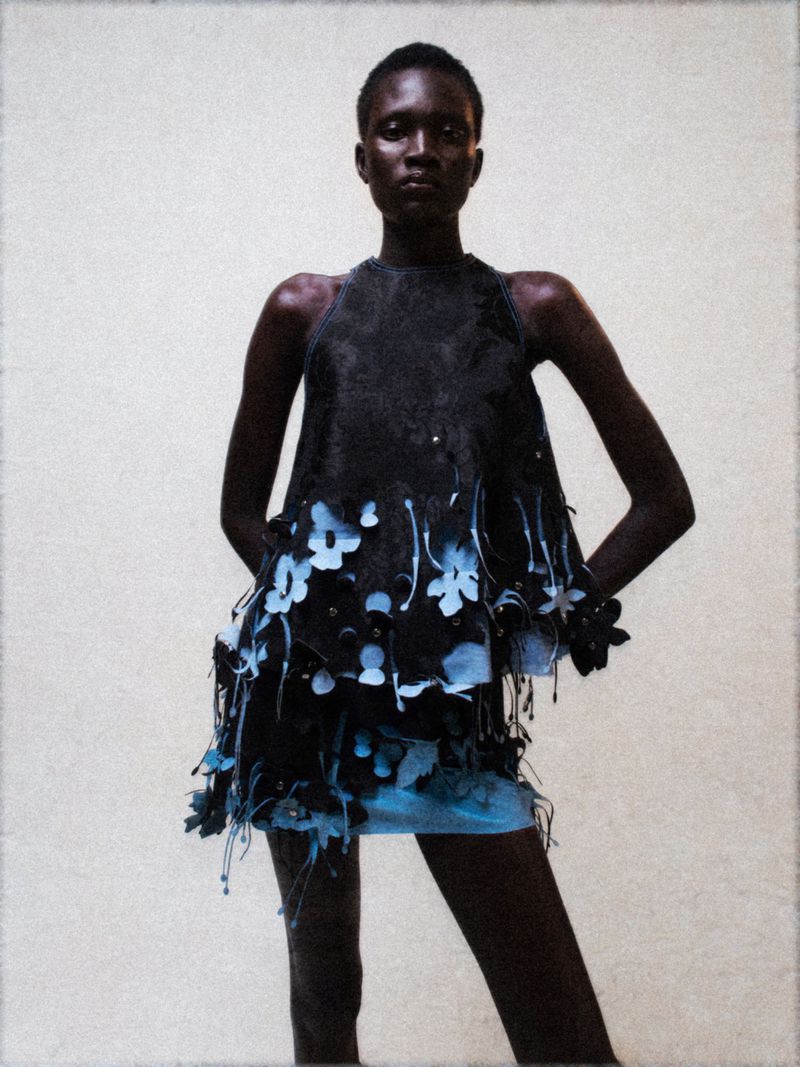 Dark-skinned woman in a floral dress poses in a studio. Fashion photography featuring clothing with floral accents and neutral background.